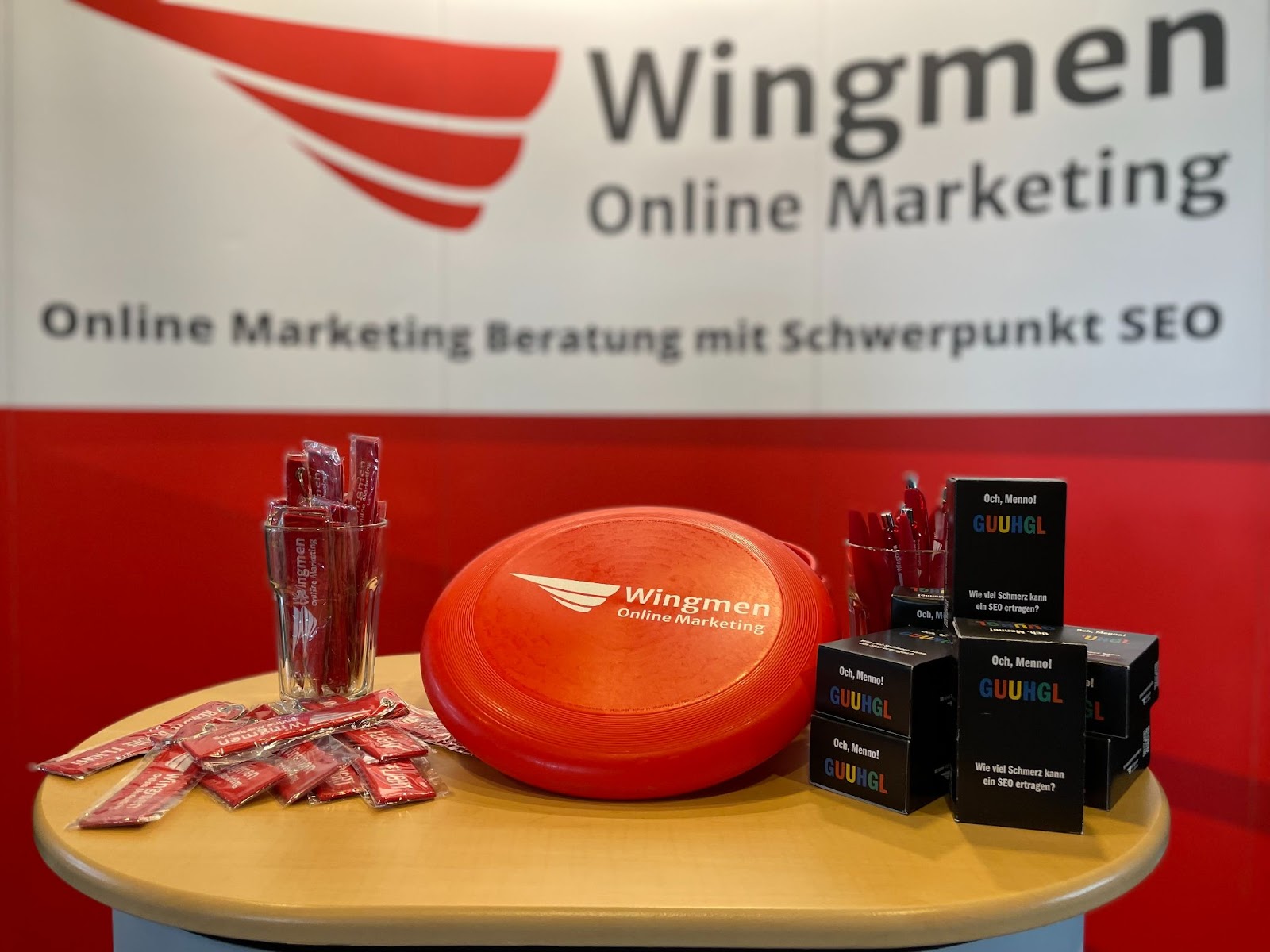 Messestand von Wingmen Online Marketing. Im Vordergrund und scharf ein kleiner Tisch auf dem Merchandise Artikel liegen: Schlüsselanhänger, Frisbees, Kullis und unser Kartenspiel. Der Hintergrund ist etwas unscharf, man kann aber trotzdem das Logo erkennen und den Schriftzug "Online Marketing Beratung mit Schwerpunkt SEO"