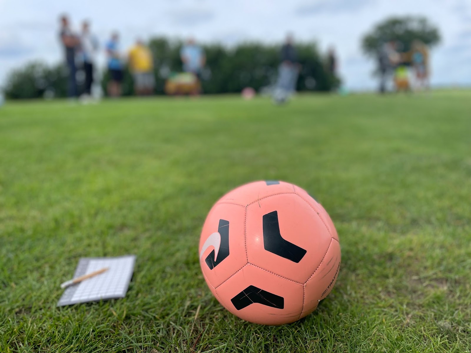 Foto vom Fußball-Golf am letzten Sonntag: Im Vordergrund liegt ein rosafarbener Fußball mit schwarzem Muster von Nike, daneben ein Zettel für die Wertung mit Stift auf dem Rasen. Im Hintergrund sind unscharf ein paar Menschen (Wingmenschen und Winglumni um genau zu sein) zu sehen.