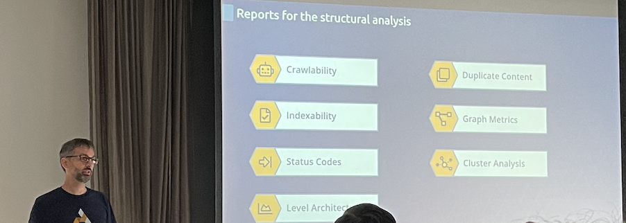 Tobi Schwarz steht neben einer Leinwand. Auf der Leinwand steht eine Aufzählungen von Reportings für die strukturelle Analyse einer Website: Crawlability, Indexability, Status Codes, Ebenenarchtitektur, Duplicate Content, Graph Metrics und Clusteranalysen.