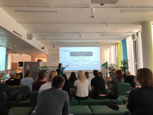 Foto vom SEO Meetup Hamburg im Juli 2019 bei XING, zu sehen ist Anke Probst beim Vortrag über Sitemaps. Das Bild ist vom anderen Ende des Raums aufgenommen, so dass die aufmerksam zuhörenden Gäste von hinten zu sehen sind. Ihr Blick ist auf Anke gerichtet, die gerade mit ihrer Hand auf dem Beamer-Bild etwas zeigt.