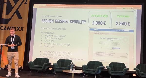 Manuel Gerlach auf der Campixx Bühne: Der Slide im Hintergrund zeigt bei Manuels Vergleich für SEObility 2.080 € CPC zu 2.940 € echter Wert